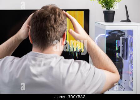 Back view of a man with hands on head, expressing distress looking at a severely broken computer screen showing colorful glitches and a modern PC setu Stock Photo