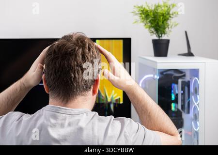 Back view of a frustrated man holding his head, looking at a broken computer monitor with a glitched display, highlighting digital issues and urgent t Stock Photo
