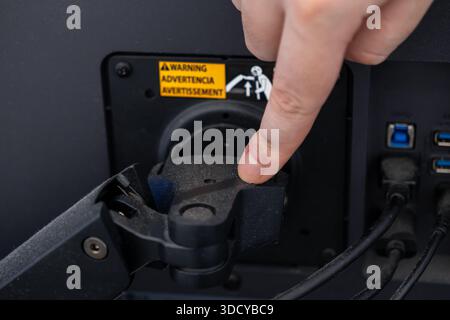 Close-up of a human finger pressing the release mechanism on a dusty computer monitor's backside, showing cable connections and a warning notice durin Stock Photo