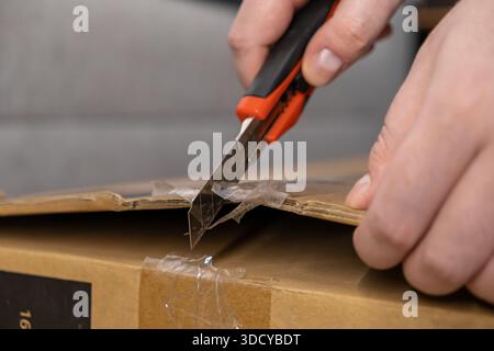 Close-up of hands using a sharp box cutter to open a taped-up brown carton, conveying the excitement of receiving an online order and starting the unb Stock Photo
