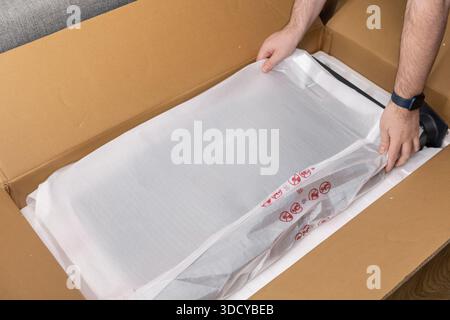 Hands carefully remove a new flat-screen display monitor wrapped in protective plastic from a large open cardboard box, illustrating product unboxing Stock Photo