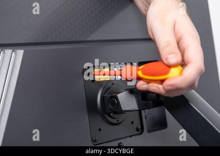 A close-up of a hand with a red and yellow screwdriver securing a mounting screw on a black monitor, demonstrating precise technology installation and Stock Photo