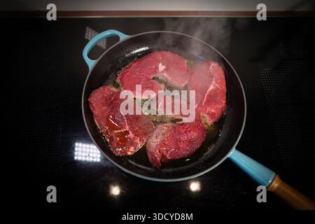 High-quality food photography showing raw beef steaks frying in a cast iron skillet on a modern stovetop. The sizzling meat, hot oil, and rising steam Stock Photo