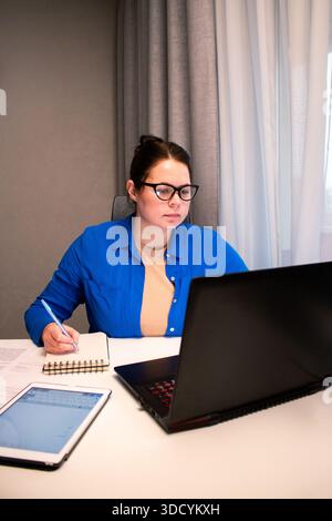 Student attending virtual university class via video conference. Young woman actively participates in online lecture, taking notes on laptop. Distance Stock Photo