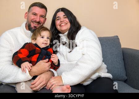 Smiling family sitting together on a couch, holding a toddler in a cozy indoor setting. Stock Photo