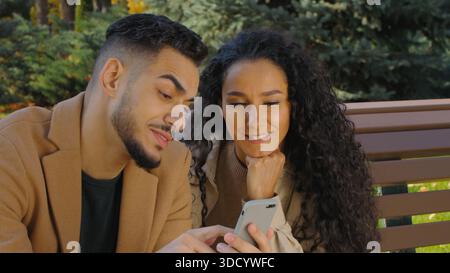 Multiethnic hispanic cheerful couple using mobile phone together in autumn city park on bench looking in screen choosing purchase ordering food Stock Photo
