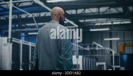 IT Manager Checking Systems in Modern Facility. Manager Walks And Monitors Safety of Modern Industrial or Data Center Facility. Supervision, Security Protocols, and Operational Flow. Back View. Stock Photo