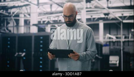IT Professional Inspecting Racks in Data Center. Bearded Male Inspecting or Troubleshooting Equipment With Handheld Device Amidst Rows Data Servers. Concept Critical Maintenance and System Management. Stock Photo