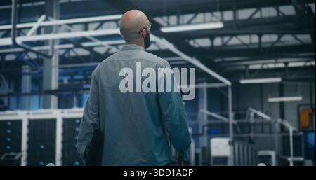 IT Manager Checking Systems in Modern Facility. Manager Walks And Monitors Safety of Modern Industrial or Data Center Facility. Supervision, Security Protocols, and Operational Flow. Back View. Stock Photo