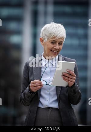 A woman with short grey hair stands outside in business attire. She holds a tablet and appears to be reflecting or planning something. Stock Photo