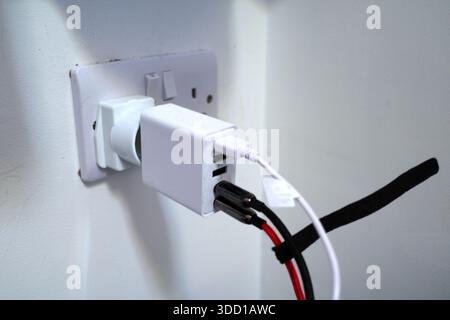A white electrical adapter is plugged into a wall socket, connected through a power converter. Three USB ports on the adapter are in use with attached Stock Photo