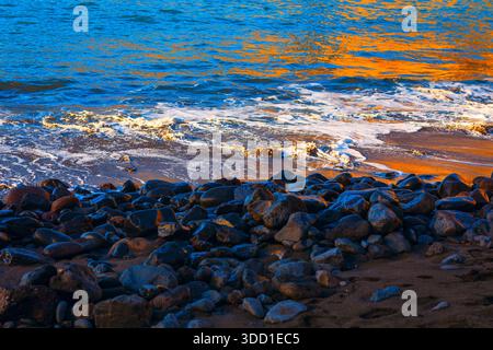 Serene moment where golden light reflects on gentle ocean waves as they meet shore covered in dark, smooth stones. Contrast between bright blue water Stock Photo