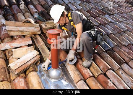 Professional roofer on a residential clay tile roof, wearing a safety harness while installing a coaxial boiler flue and applying waterproof sealing t Stock Photo