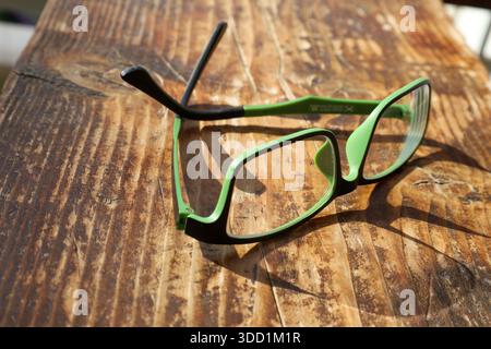 A fashionable pair of green and black eyeglasses rests on a rustic wooden table, lit by warm afternoon sun. Stock Photo