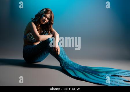 A woman poses elegantly in a mermaid costume with intricate body art, showcasing her vibrant tail and accessories. || Model released Stock Photo