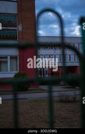 Soviet era school in Minsk is seen through green fence bars, red brick and pale tiles, rows of windows, Russian welcome sign, overcast clouds, late se Stock Photo