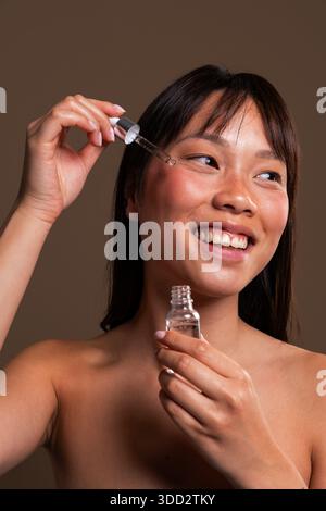 Beautiful chinese woman portrait on colorful background. Asian woman smiling, applying cosmetic serum to her cheek with a dropper. Focusing on beauty, Stock Photo
