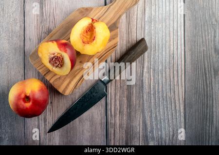 Top-down view of two halved nectarines, one whole fruit, and a chef's knife on a natural olive wood cutting board set against a rustic wooden backgrou Stock Photo