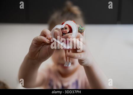 Child holding small toy doll inside heart shaped plastic frame, showing childhood play, imagination, and early development through simple toys at home Stock Photo