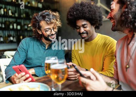 Three diverse young adult friends using mobile phone sitting on table at bar Stock Photo