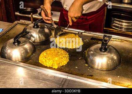Two hands shape round okonomiyaki patties with spatulas on the teppan griddle of a restaurant in Osaka, Japan, while three other portions cook under metal domes nearby. Okonomiyaki means "grilled as you like it": a batter of wheat flour, egg and dashi is mixed with finely shredded cabbage and cooked on the hot plate. The dish is regarded as Osaka's signature food. Stock Photo