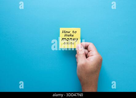 Hand holding a yellow sticky note with how to make money online handwritten on a blue background Stock Photo