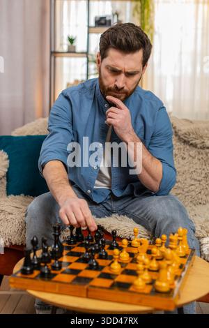 Young man sitting at home on sofa, playing chess alone, making a serious move with deep concentration. Caucasian guy strategizing, feeling thoughtful Stock Photo
