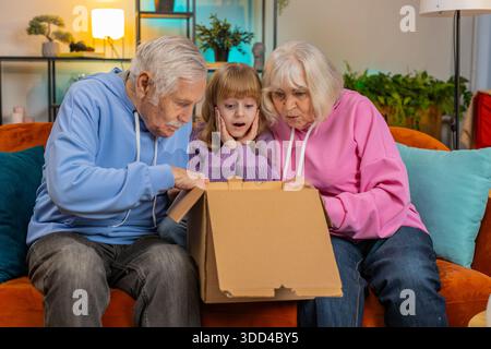 Happy excited Caucasian grandparents and granddaughter girl opening cardboard box together sits on sofa at home. Smiling family consumers unpack good Stock Photo