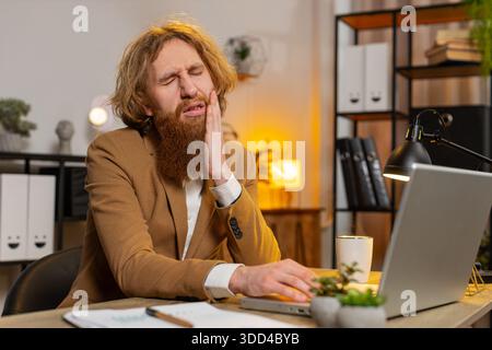 Caucasian businessman freelancer working on laptop and feeling toothache touching mouth with hand, painful expression because of dental illness. Caries, pulpitis, gum disease, dental disease concepts. Stock Photo
