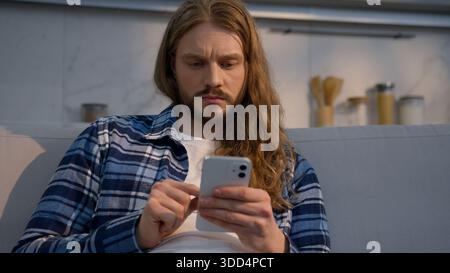 Happy smiling Caucasian man sitting on couch at home using mobile phone indoor male relaxed carefree guy scrolling social media apps smartphone Stock Photo