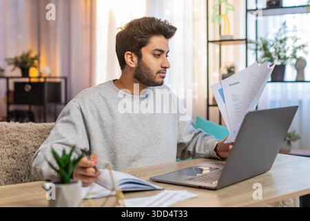 Young Indian man freelancer sitting at table reviewing financial reports on laptop, holding documents and receipts, taking notes in notebook. Hispanic guy sits nearby at home, focused on his own task. Stock Photo