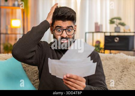 Indian man at home checks bills receipts with tense expression, nervously grabbing his head in frustration. Arabian guy on sofa struggles with financial pressure and visible signs of emotional stress Stock Photo