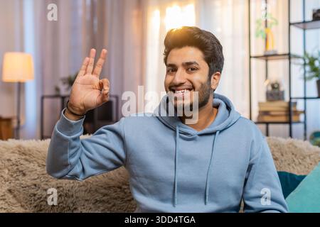 Happy cheerful Indian man looking approvingly at camera, showing ok gesture, positive like sign. Arabian Hindu guy sitting on sofa in modern apartment Stock Photo