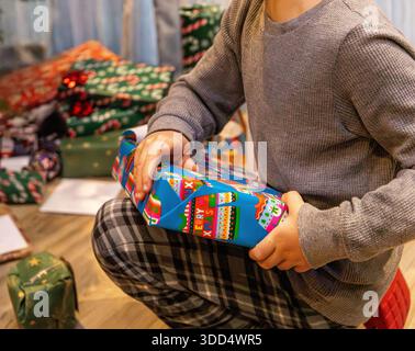 Child holding a wrapped Christmas present while kneeling on the floor surrounded by other festive gifts Stock Photo