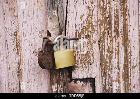 Two padlocks, one rusty vintage and one new brass, secured on a weathered pink wooden door. Concept of security, age contrast, and rustic texture. Stock Photo
