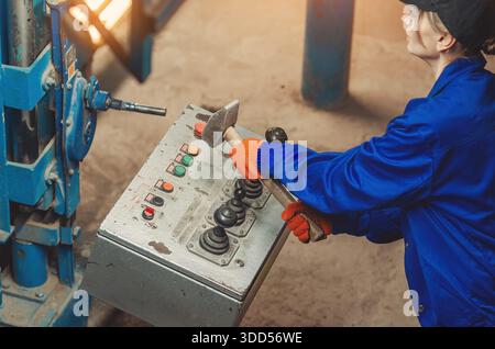 Female engineer in blue suit and protective gloves working with control panel, monitoring and automating industrial processes. Hammer in hands as conc Stock Photo