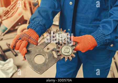 Close-up view of worker s hands in orange protective gloves holding metal gears in industrial workshop. Focus on machinery parts, craftsmanship, and m Stock Photo