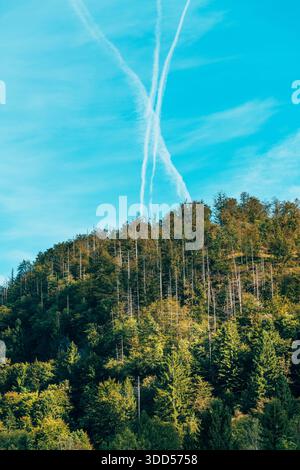 Crossing jet trails forming X shape over woodland in alpine mountain landscape. Selective focus. Stock Photo
