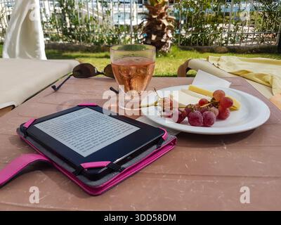 A plate of cheese and grapes, alongside a glass of rosé wine, an Amazon Kindle e-reader and a pair of sunglasses, sit on a table outside a hotel in Tu Stock Photo