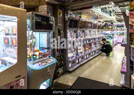 A man leans over a crane-game machine filled with plush toys in an arcade store in Osaka, Japan. A single spin of a gachapon machine typically costs 200 to 500 yen, payable only in cash coins. Stock Photo
