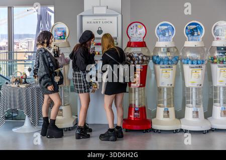 Three young women in the Umeda Sky Building, Osaka, at a vending machine for heart locks. Heart locks are very popular in Japan, alongside the ubiquitous gachapon vending machines. Three young women are shopping at a vending machine shop in the Umeda Sky Building. Osaka, Osaka Prefecture, Japan Stock Photo