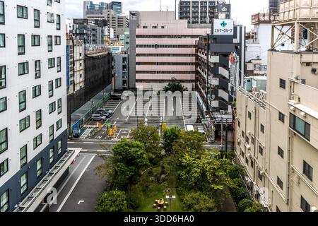 A small green park with trees sits wedged between a car park and multi-storey residential and office buildings. Osaka, Japan. Stock Photo