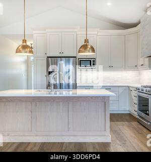 This bright, contemporary residential kitchen showcases a refined balance of modern design, functionality, and understated luxury. Dominated by a clean white palette, the space features custom cabinetry, seamlessly integrated stainless steel appliances, and a large central island that serves as both a practical workspace and a social hub. The expansive island is topped with a sleek, light-toned stone or quartz countertop, emphasizing durability and elegance while providing generous surface area for food preparation, entertaining, or casual dining. Warm brass pendant lights hang above. Stock Photo