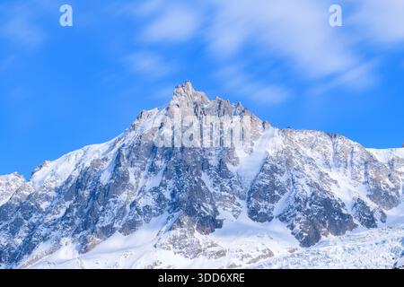 The dramatic, snow-covered summit of Aiguille du Midi rises sharply against a vivid blue sky, with soft, fast-moving clouds adding a sense of motion above rugged alpine slopes. Stock Photo