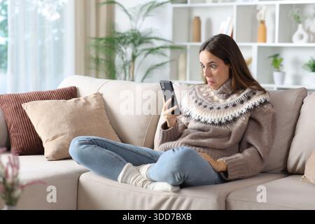 Perplexed woman in winter checking phone sitting on a sofa at home Stock Photo