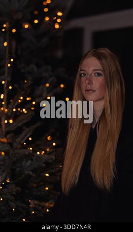 This is a portrait photograph of a young woman set against the backdrop of a Christmas tree illuminated with warm-toned string lights. The woman has l Stock Photo