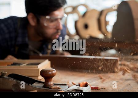 Close-up of a hand plane on a workbench with wood shavings flying as a carpenter blows away dust. Workshop tools and blurred craftsman in background, Stock Photo
