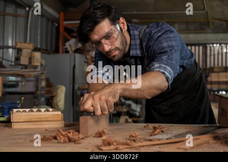 Close-up of a focused carpenter wearing safety goggles using a hand plane on wood at a workbench. Wood shavings and tools in a workshop, concept of cr Stock Photo