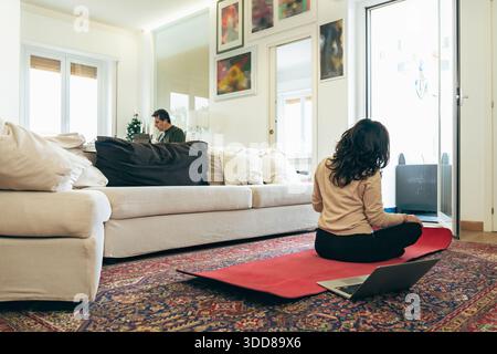 Middle aged couple at home sharing space while balancing remote work and personal wellness. Woman practicing yoga on mat while man works on laptop in Stock Photo