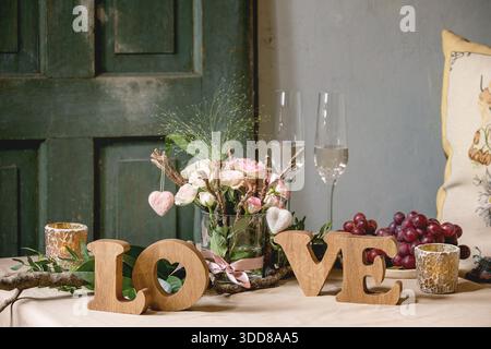 st. Valentines day or wedding romantic table setting with wooden letters love, needle felted hearts, bouquet of pink roses, couple glasses of champagne and red grapes on natural linen tablecloth. Stock Photo